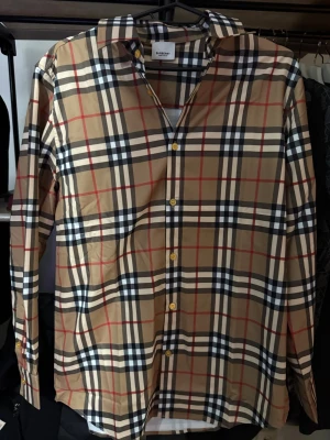 Rutig Burberry skjorta i bomull - Klassisk rutig skjorta från Burberry i beige, svart, vit och röd. Skjortan har långa ärmar, knappar framtill och ikoniskt rutmönster. Tillverkad i mjuk bomull och har snygga detaljer som guldfärgade knappar vid kragen.