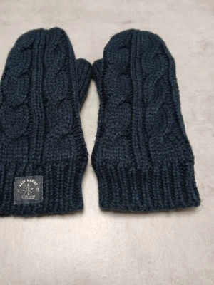 Mörkblå stickade Race Marine-vantar - Ett par stickade tumvantar med lös mudd i öppningen. Jag tror att insidan är fleece. Längd: cirka 25 cm Bredd: cirka 11 cm Yttertyg: 100 % akryl Foder: 100 % polyester Se bilder för skick och passform. Färgen på bilderna, speciellt närbilderna, stämmer inte alltid överens med verkligheten.