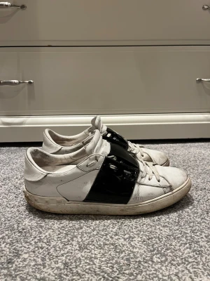 Valentino Garavani Open sneakers vit/svart Strlk 44 - Säljer ett par Valentino Garavani Open sneakers i vitt skinn med bred svart lackdetalj över snörningen. Klassisk rund tå, platt sula och diskret logga på plösen. Snygg kontrast mellan det vita och svarta, perfekt för dig som gillar statement-sneakers. Nypris: 7280kr, mitt pris 1499kr. Pris kan diskuteras!