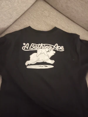 Svart BAPE x Coca-Cola t-shirt med isbjörnar - Svart t-shirt från A Bathing Ape i samarbete med Coca-Cola. Stor tryck på framsidan med isbjörnar och BAPE-logga i blått och vitt, samt Coca-Cola-flaskor. Baksidan har stor vit BAPE- och isbjörnsgrafik. Klassisk passform och crewneck.