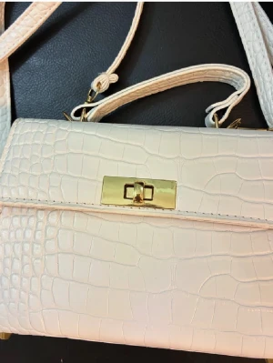 Vit axelväska med krokopräglad yta - Snygg vit axelväska med krokopräglad yta och guldfärgade metalldetaljer. Väskan har ett stilrent, rektangulärt format med praktiskt handtag och axelrem. Stängs med ett elegant guldfärgat lås framtill. Perfekt för dig som gillar clean och lyxig vibe.