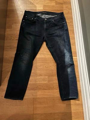 Mörkblå jeans från Nudie Jeans - Snygga mörkblå jeans från Nudie Jeans med klassisk femficksdesign och subtila slitningar. Jeansen har raka ben och normal passform, med Nudie-logga på bakfickan. Materialet är robust denim i bomull och färgen har en cool tvättad look. Modellen är lean Dean storlek w34 l30