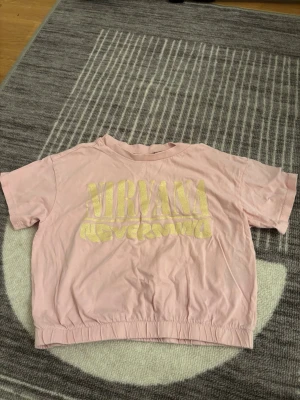 Rosa Nirvana Nevermind t-shirt - Säljer en rosa t-shirt med guldfärgat Nirvana Nevermind-tryck på bröstet. T-shirten har korta ärmar och en croppad passform med resår i nederkant. Perfekt för dig som gillar bandtröjor och vill sticka ut med en cool färg.