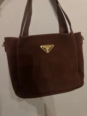 Snygg brun axelväska i mjuk mocka med klassisk trekantig logga i guld på framsidan. Väskan har stilrena sömmar och rymligt fack med ljusbrunt innerfoder. Perfekt storlek för att få plats med allt du behöver och har guldfärgade detaljer.
