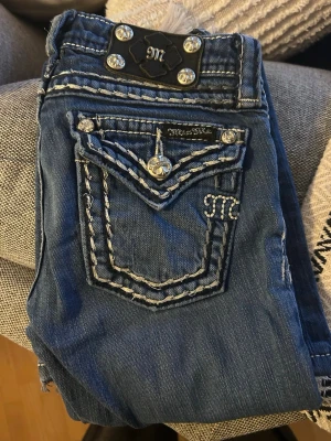 Miss Me jeans W23 - Snygga blå jeans från Miss Me med kontrasterande vita sömmar och broderade detaljer på fickorna. Dekorativa knappar med blommönster och logotyp på bakfickan. Klassisk femficksmodell med dragkedja och knappstängning. Perfekt för dig som gillar statement-jeans.