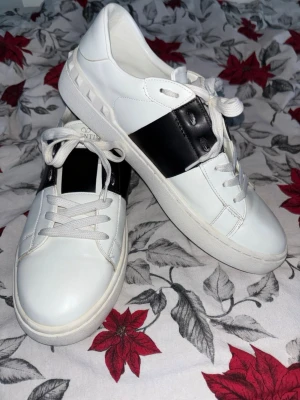 Valentino Garavani Open sneakers - Snygga Valentino Garavani Open sneakers i vitt skinn med bred svart läderdetalj över snörningen. Klassisk rund tå, platt sula och coola nitar längs hälen. Perfekt för dig som gillar lyxiga och stilrena sneakers med unik design.