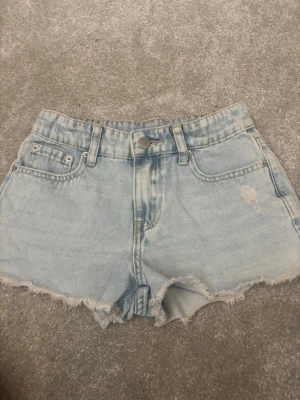 Ljusblå denimshorts från UNIQ - Säljer ett par ljusblå denimshorts från UNIQ med fransig kant och slitna detaljer. Klassisk femficksmodell med knapp och dragkedja framtill. Perfekta för sommaren och ger en avslappnad vibe.