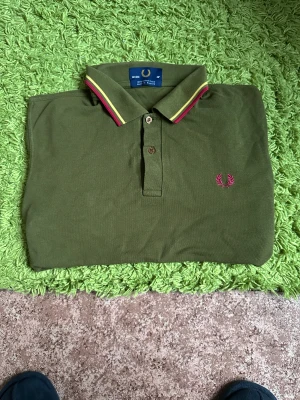 Olivgrön Fred Perry pikétröja - Säljer en olivgrön pikétröja från Fred Perry med klassisk krage och knäppning framtill. Kragen har gula och röda ränder och bröstet pryds av Fred Perrys ikoniska rosa lagerkrans. Tröjan är tillverkad i 100% bomull och har en tidlös stil.