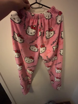 Hello Kitty rosa mjukisbyxor - Supergulliga rosa mjukisbyxor med Hello Kitty-tryck över hela byxorna. De har elastisk midja och är gjorda i mjukt fleece-material, perfekta för chill hemma eller sleepover. Söt design med Hello Kittys ansikte och röda rosett.