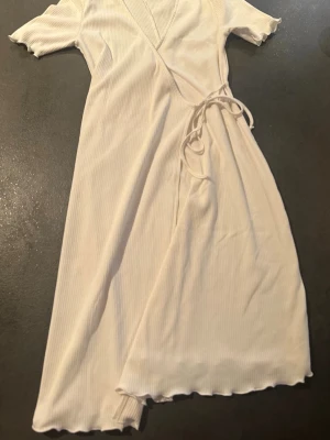 Vit omlottklänning från Primark - Supersöt vit omlottklänning från Primark med korta ärmar och ribbad struktur. Klänningen har knytband i midjan och vågiga kanter vid ärmslut och nederkant. Perfekt för alla tillfällen och ger en fräsch och enkel look.