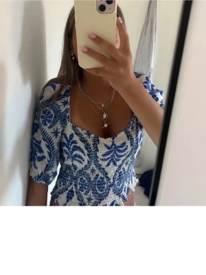 Blåvit croppad blus med mönster - Säljer en croppad blus i blått och vitt med tropiskt blommigt mönster. Toppen har puffiga korta ärmar, fyrkantig ringning och smockad midja med volangkant. Perfekt för dig som gillar somriga vibes och snygga detaljer.