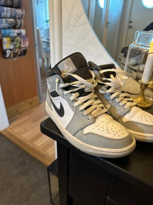 Nike Air Jordan 1 Mid - Säljer ett par Nike Air Jordan 1 Mid i grått och vitt med svarta detaljer och klassisk svart swoosh. Skorna har snörning, perforerad tåbox och hög siluett. Tillverkade i skinn och syntetmaterial för en clean och tidlös look. Perfekta för dig som gillar streetwear.