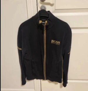  ziptröja Hugo boss - Snygg svart ziptröja från BOSS med guldfärgade detaljer och dragkedja. Tröjan har hög krage, fickor framtill och BOSS-logga broderad på bröstet. Perfekt för dig som vill ha en stilren och exklusiv look.