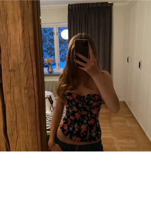 Svart blommig korsett med färgglada rosor - Säljer en snygg korsett med svart botten och blommigt mönster i rosa, rött, blått och grönt. Toppen är axelbandslös och har en figurnära passform som framhäver midjan. Perfekt för dig som vill sticka ut med färg och mönster!