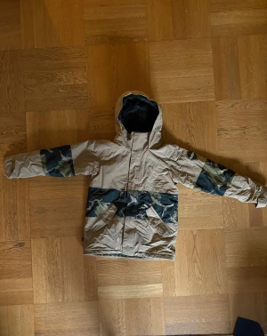 Beige Burton vinterjacka med camo detaljer - Beige Burton vinterjacka med camo detaljer storlek 163 för barn 