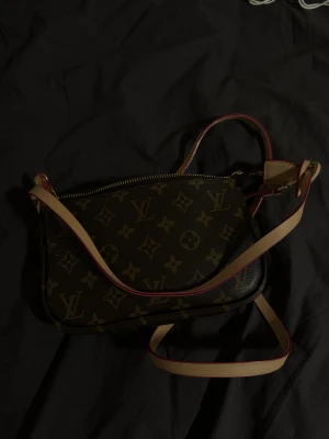 Brun axelväska - Snygg axelväska i klassiskt monogrammönster. Väskan är brun med beige och guldiga detaljer, har justerbar axelrem i ljust skinn och dragkedja upptill. Köpt i Spanien 