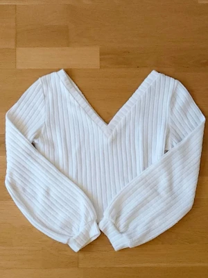 Vit tröja - Vit ribbad tröja med v-ringning och långa ärmar. Tröjan har en croppad passform och breda muddar vid ärmsluten. Perfekt för dig som gillar en enkel men ändå snygg stil. Materialet känns mjukt och stretchigt.