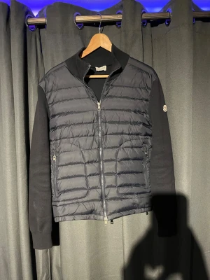 Mörkblå Moncler cardigan - Svart pufferjacka från Moncler med quiltad framsida och stickade ärmar. Den är storlek medium men sitter som Small. Jackan har dragkedja framtill, två sidofickor med dragkedja och Moncler-logga på vänster ärm. Materialet är en mix av dun och stickad textil, perfekt för kyliga dagar.