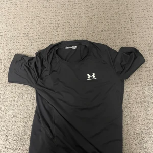Svart Under Armour HeatGear t-shirt XL - Svart Under Armour HeatGear t-shirt i storlek XL med tryckt logga på bröstet. T-shirten är kortärmad och har en tight passform, perfekt för träning. Materialet är polyester som andas och håller dig sval under intensiva pass.