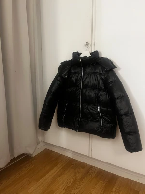 Svart pufferjacka från lager 157, storlek S - Svart pufferjacka från lager 157 med glansig finish och hög krage. Jackan har två sidofickor med dragkedja, quiltad design och en luva. Perfekt för kalla dagar och har en snygg, modern look som funkar till streetwear💞.
