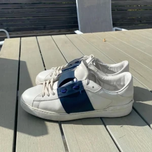Valentino Open sneakers vit/blå - Säljer ett par Valentino Open sneakers i vitt skinn med bred marinblå läderpanel över mitten och blå detaljer vid snörningen. Klassisk rund tå och platt sula, med diskret Valentino-logga på tungan. Snygg och stilren design som sticker ut. Storlek 44 men passar jättebra både 43 och 45. Nypris: 3499kr