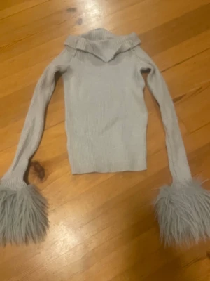 Ljusgrå stickad tröja med pälsärmar - Unik ljusgrå stickad tröja med långärmade ärmar som avslutas med stora fluffiga pälsdetaljer. Tröjan har en ribbad struktur och en snygg krage som ger extra stilpoäng. Perfekt för dig som vill sticka ut och ha något annorlunda i garderoben.