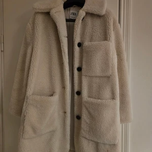 Beige teddyjacka från Zara - Säljer en beige teddyjacka från Zara i storlek S. Jackan har stora fickor framtill, krage och svarta knappar. Den är lång och har en mysig, fluffig känsla som passar perfekt när det är kallt ute. Perfekt för dig som gillar en chill och trendig look.
