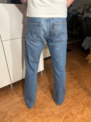 Levi's 501 blå jeans straight fit - Klassiska Levi's 501 jeans i blå denim med straight fit och normal passform. Byxorna har fem fickor, knappgylf och den ikoniska röda Levi's-taggen på bakfickan. Perfekt för dig som gillar tidlös stil och äkta jeanskänsla. Modellen är 190 cm lång och väger 70 kg.