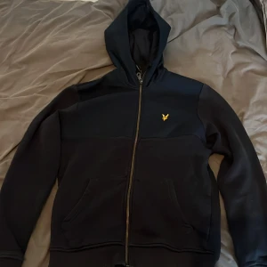 Svart huvtröja från Lyle & Scott - Säljer en svart huvtröja från Lyle & Scott med dragkedja och en liten gul logga på bröstet. Tröjan har långa ärmar och en praktisk huva. Perfekt för en avslappnad stil.