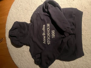 Svart hoodie Acne Studios Stockholm - Svart hoodie från Acne Studios med tryck 'Stockholm 1996' på baksidan. Tröjan har huva, ribbade muddar och är tillverkad i mjuk bomull med inslag av polyester. Perfekt för dig som gillar stilrena och klassiska streetwear-plagg. Sitter ungefär en storlek större så om du har S i vanliga fall passar den perfekt 🤩 