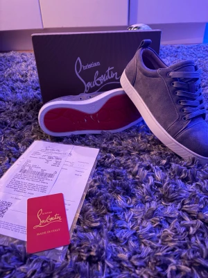 Grå Christian Louboutin sneakers - Snygga grå sneakers från Christian Louboutin med klassisk röd undersida och vit kant. Skorna har rund tå, snörning och är tillverkade i mocka med skinnfoder. Perfekt för dig som vill ha en lyxig och stilren look med exklusiv känsla.