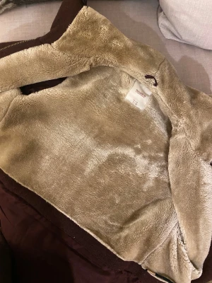 Vinröd jacka från Pull&Bear - Säljer en vinröd, långärmad parkajacka från Pull&Bear med mjukt beige teddyfoder på insidan. Jackan har hög krage med knappar, dragkedja och fickor med dragkedja. Perfekt för kalla dagar.