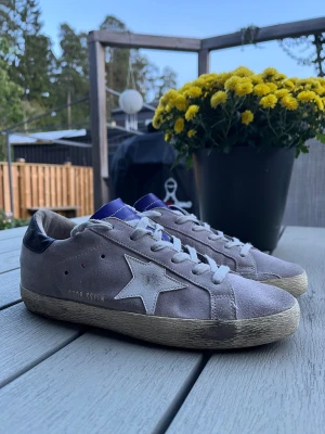 Grå sneakers från Golden Goose - Säljer ett par gråa Golden Goose sneakers med vit stjärndetalj på sidan, lila plös och svart häl. Skorna har snörning och är tillverkade i mocka med platt sula och rund tå. Perfekt för dig som gillar en cool och avslappnad stil.