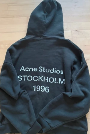 Svart hoodie Acne Studios Stockholm - Svart hoodie från Acne Studios med stor huva och tryck på ryggen: 'Acne Studios STOCKHOLM 1996'. Tröjan har lång ärm och klassisk loose passform. Perfekt för dig som gillar streetwear och enkel stil.