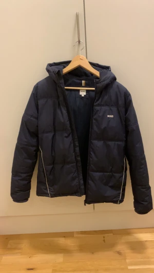 Boss Vinterjacka  - Snygg mörkblå vinterjacka från BOSS med huva. Håller värmen extremt bra nu när det är som kallast. Jackan har dragkedja framtill och vita detaljer längs sidorna. Materialet är polyester och vadd och är perfekt nu i för vintern  Nypris ligger på 2000kr Mitt pris 1100kr men pris kan diskuteras 