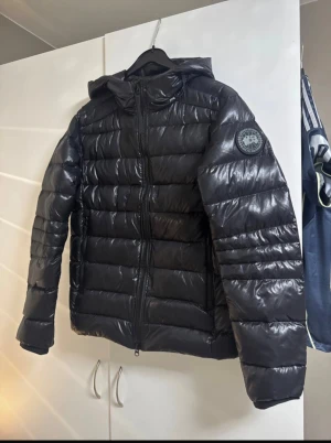 Canada goose jacka  - Säljer en svart pufferjacka från Canada Goose med glansig finish och huva. Jackan har en tydlig logotyp på ärmen och dragkedja framtill med Canada Goose-tryck. Perfekt för kalla dagar och har en modern, clean look.