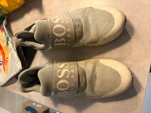Grå sneakers från Hugo Boss - Snygga grå sneakers från Hugo Boss med bred resår över foten där det står BOSS i stora bokstäver. Ovandelen är i mesh och syntetmaterial för en sportig look. Skorna har rund tå och platt sula, perfekta för en clean och modern stil.