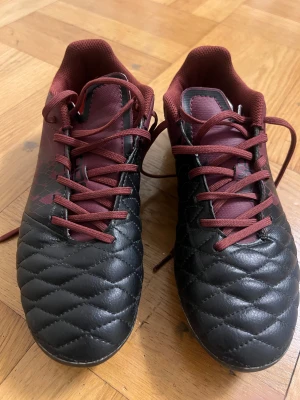 Svarta och vinröda fotbollsskor Decathlon - Säljer ett par fotbollsskor från Decathlon i storlek 39. Skorna är svarta och vinröda med snörning och mönstrad ovandel. Utrustade med fasta dobbar för bra grepp på planen. Ovandelen är i syntetmaterial och insidan är vadderad för extra komfort.