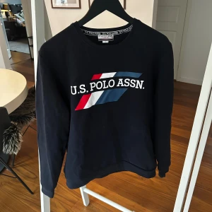 Mörkblå sweatshirt från U.S. Polo Assn. - Snygg mörkblå sweatshirt från U.S. Polo Assn. med stort tryck på bröstet i vitt, rött och blått. Tröjan har rund hals och långa ärmar, perfekt för en chill och sportig look. Materialet är mjuk bomull och passformen är normal.