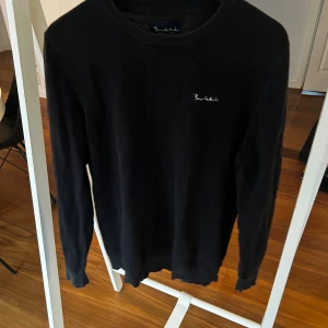 Svart stickad tröja från Bondelid - Svart stickad tröja från Bondelid i storlek L. Klassisk rund halsringning och diskret broderad logga på bröstet. Mjuk och skön kvalitet som passar perfekt till jeans eller chinos. Långärmad modell med ribbade muddar vid ärmslut och nederkant.