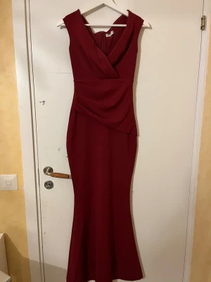 Vinröd långklänning från Goddiva - Säljer en elegant vinröd långklänning från Goddiva London med omlott-detalj framtill och v-ringad hals. Klänningen är ärmlös och har en figurnära passform som framhäver siluetten. Materialet är mjukt och har en lätt stretch. Perfekt för bal eller fest! Klänningen är upp sydd lite men inget som syns 