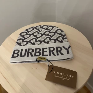 Vit och grå Burberry mössa - Snygg vit och grå mössa från Burberry med stort logotyp-tryck framtill och grafiskt mönster i form av hjärtan. Mössan är stickad och har en uppvikt kant. Perfekt accessoar för att lyfta din vinterstil.