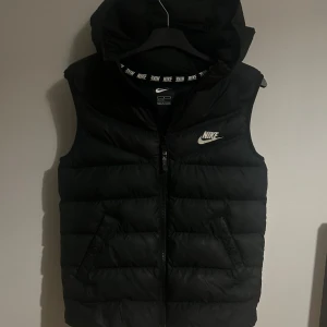 Svart dunväst från Nike med huva - Priset kan diskuteras!                                                      Svart dunväst från Nike med huva och dragkedja framtill. Västen har två snedställda fickor med knappstängning och ett broderat Nike-logo på bröstet. Insidan har ett band med Nike-tryck runt halsen. Perfekt för lager-på-lager och streetstyle.