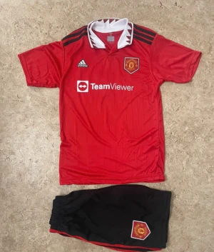 Manchester United Ronaldo 7 fotbollströja - Säljer en röd Manchester United fotbollströja från Adidas med vita och svarta detaljer, korta ärmar och klassisk krage. Tryck med TeamViewer, klubbmärke och Ronaldo 7 på ryggen. Materialet är lätt och andas, perfekt för match eller träning.