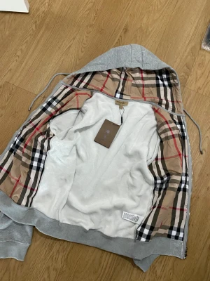 Grå Burberry hoodie med dragkedja - Säljer en grå hoodie från Burberry med klassiskt rutigt foder i huvan och insidan. Tröjan har hel dragkedja, snörning i huvan och Burberrys broderade logga på bröstet. Mjuk bomullskvalitet och två fickor framtill. Perfekt för dig som gillar stilren streetwear.