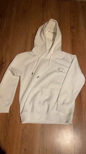 Vit hoodie från Oakley med huva - Snygg vit hoodie från Oakley med klassisk huva och snörning. Tröjan har en stor magficka och Oakleys logga broderad på bröstet. Tillverkad i mjuk bomull och har långa ärmar med ribbade muddar. Perfekt för chill dagar.