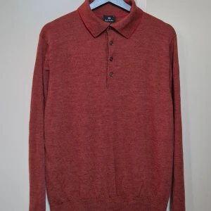 Röd merinoull pikétröja Paul Smith XL - Snygg röd stickad pikétröja från Paul Smith i storlek XL. Tröjan har klassisk krage med diskret röd kant, tre knappar framtill och broderad PS-logga vid nederkanten. Långärmad och mjuk kvalitet som känns chill och stilren. 100% merinoull.