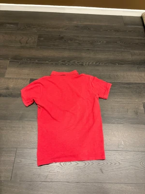 Röd piké från Polo Ralph Lauren - Snygg röd pikétröja från Polo Ralph Lauren med klassisk krage och två knappar framtill. Hör av dig vid frågor. Den sitter som XS/S 