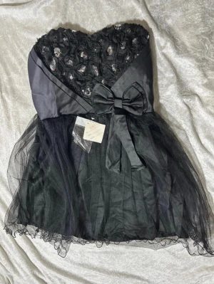 Svart kort festklänning  - Supersöt svart axelbandslös klänning med hjärtformad överdel dekorerad med blommiga applikationer och paljetter. Midjan pryds av en stor rosett och kjolen är i flera lager tyll för extra volym. Perfekt för dig som vill sticka ut med en glamorös look.