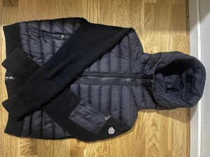 Svart Moncler pufferjacka med huva - Svart cardigan  från Moncler med stickade ärmar och rygg, quiltad framsida och huva. Jackan har tvåvägsdragkedja, ribbade muddar och Moncler-logga på ärmen. Materialmix av polyester och stickad textil ger en unik look. Perfekt för dig som gillar streetstyle. Kvitto finns. 